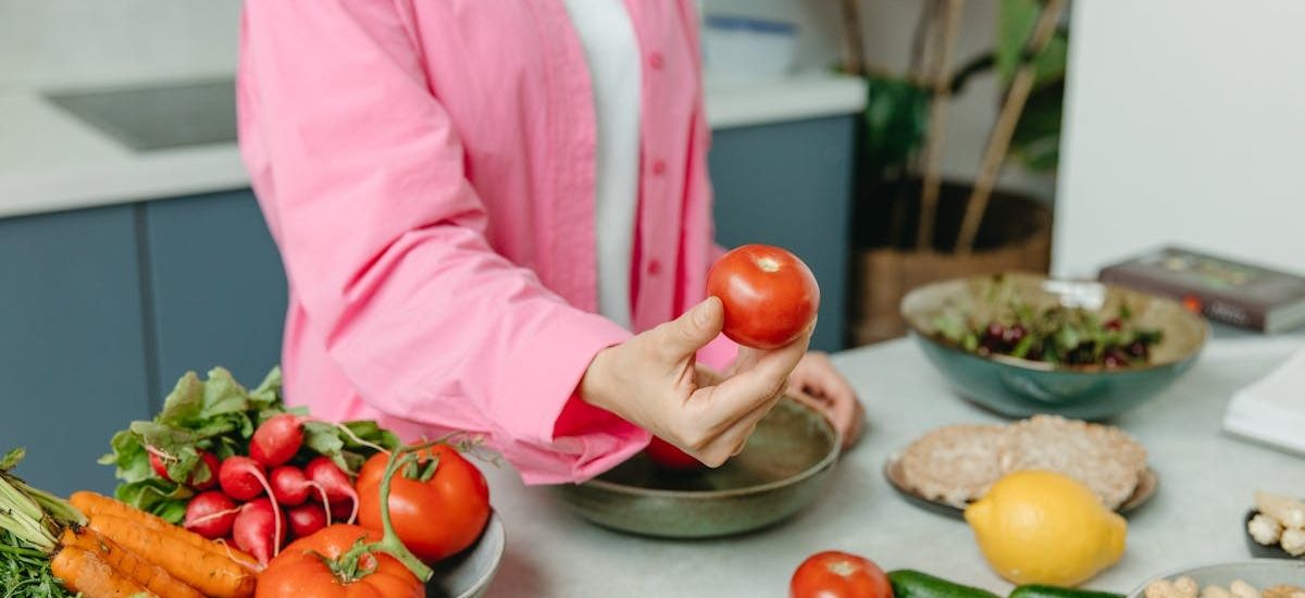 Person in pink overshirt holding a fresh tomato with various organic vegetables on a countertop.
