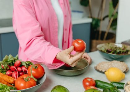 Person in pink overshirt holding a fresh tomato with various organic vegetables on a countertop.